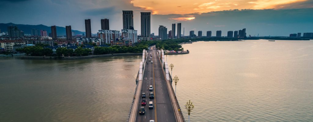 Aerial view of a long bridge over a wide body of water at sunset, with cars driving toward a city skyline filled with high-rise buildings under a dramatic, cloud-filled sky