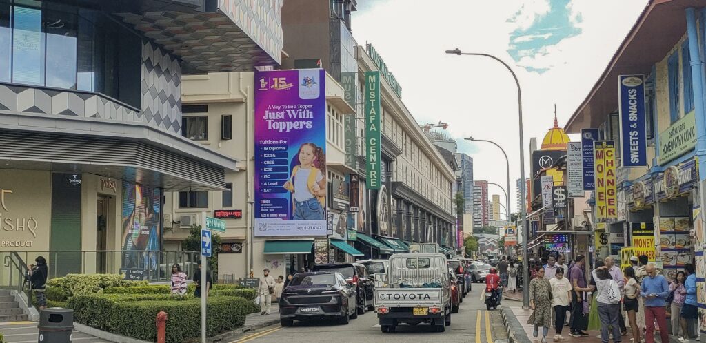 A vertical billboard near Mustafa Centre on Syed Alwi Road, Singapore, displaying an advertisement for Toppers Education tuition services. The ad features a young girl with a yellow backpack against a purple and blue gradient background, listing courses such as IB Diploma, IGCSE, CBSE, ICSE, A Level, and SAT, with busy street traffic and pedestrians in the foreground.