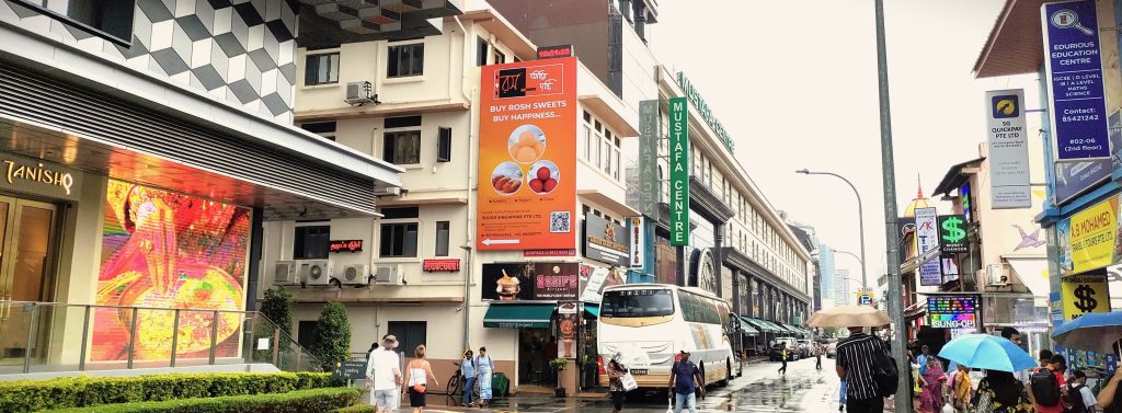 A vertical billboard near Mustafa Centre, Singapore, showing Shori Singapore’s advertisement with images of traditional Indian sweets such as rosh sweets, yogurt, and ghee on a bright orange background.