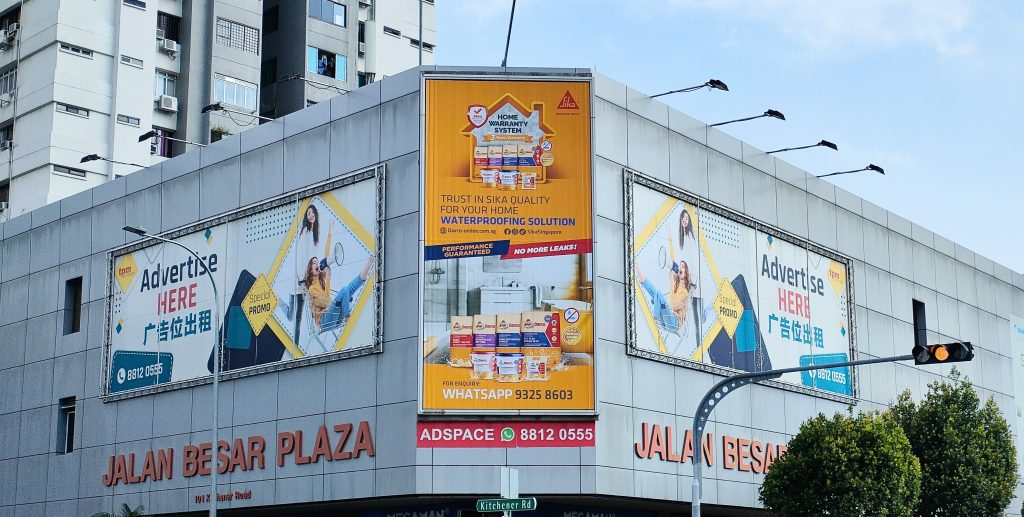 A large billboard at Jalan Besar Plaza, Singapore, featuring the Sika Home Warranty System advertisement for home waterproofing solutions, with a dominant yellow background and promotional text centered on the building facade.