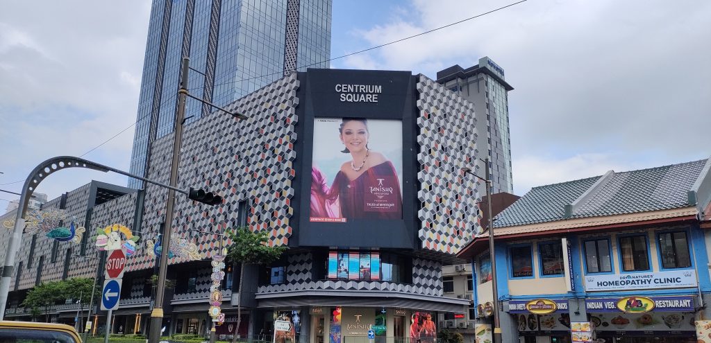 A massive LED screen at Centrium Square, Singapore, featuring a female model wearing a maroon dress and luxury jewelry in Tanishq’s Tales of Mystique campaign.