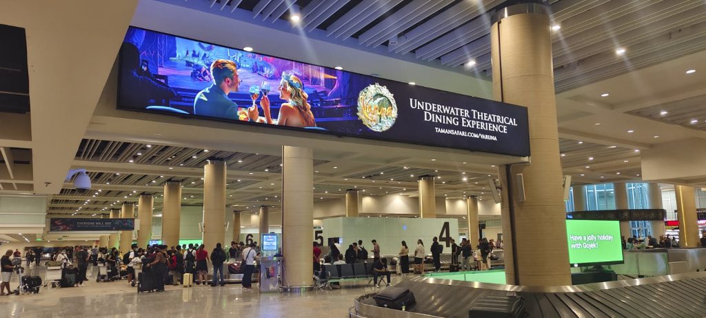 Baggage claim area inside an airport terminal with travelers waiting beside luggage carousels and large numbered pillars, featuring a wide overhead digital billboard by The Perfect Media promoting the Bali Safari underwater theatrical dining experience